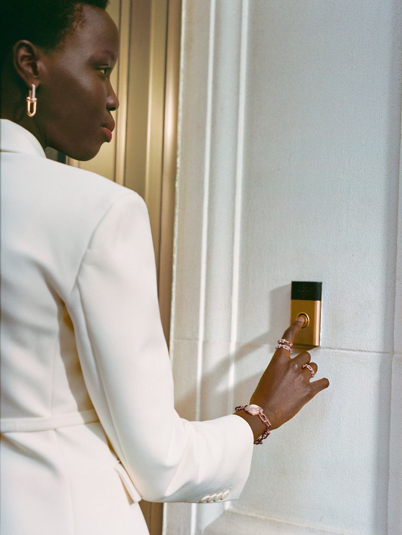 Elegant Black woman in a white suit pressing a gold doorbell, accessorized with a bracelet and rings. Indoor, minimalist, fashion shot.