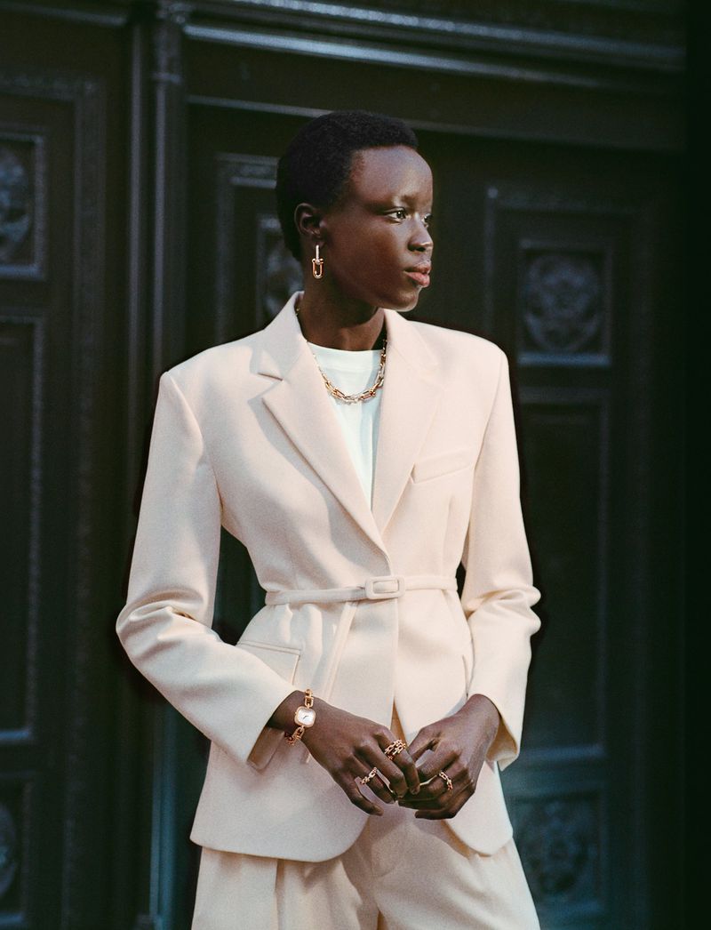 A poised Black woman in a belted beige suit accessorized with jewelry, stands against a dark textured door, radiating style and confidence.
