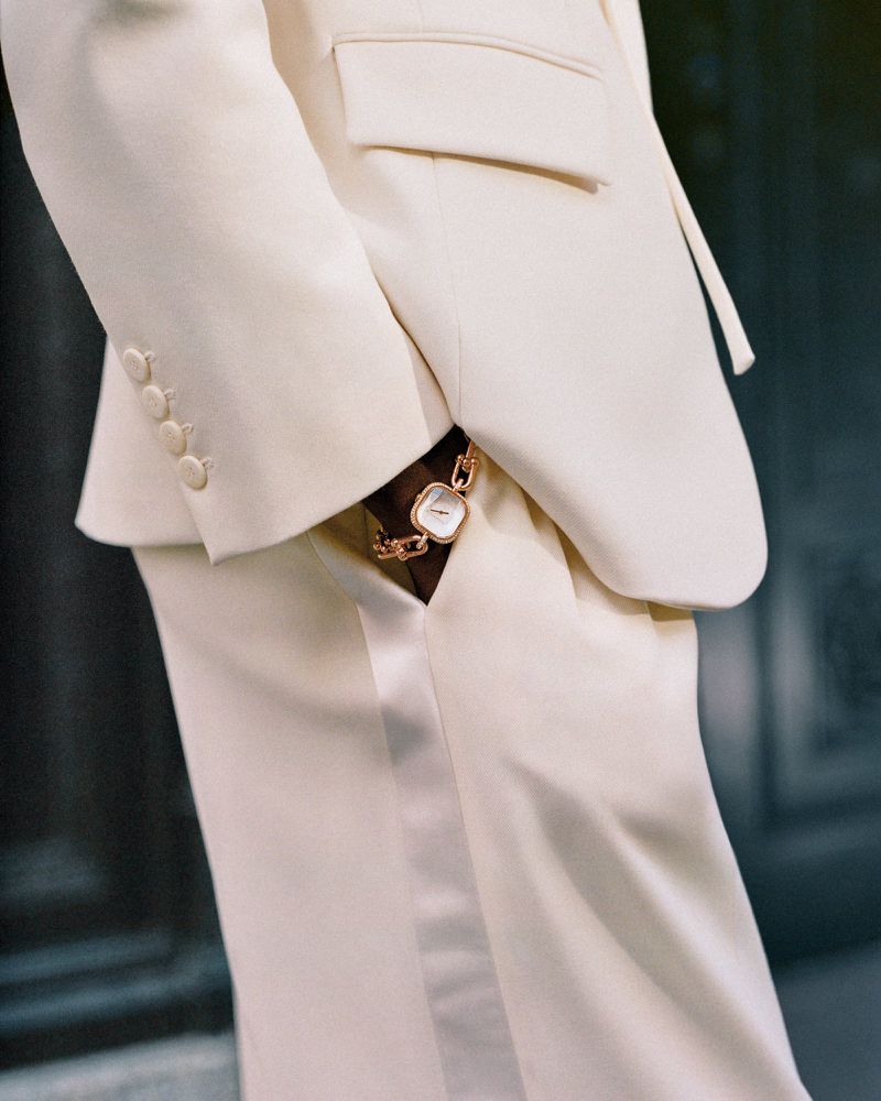 Close-up of a stylish cream-colored suit with a square-faced gold watch accentuating the wrist, set against a neutral backdrop.