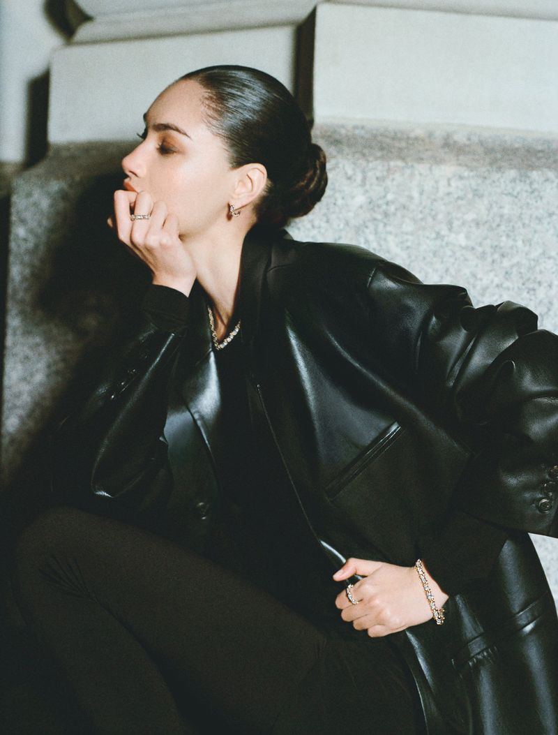 A young adult woman with slicked-back hair poses in a black leather jacket and leggings, seated on steps with a neutral expression.