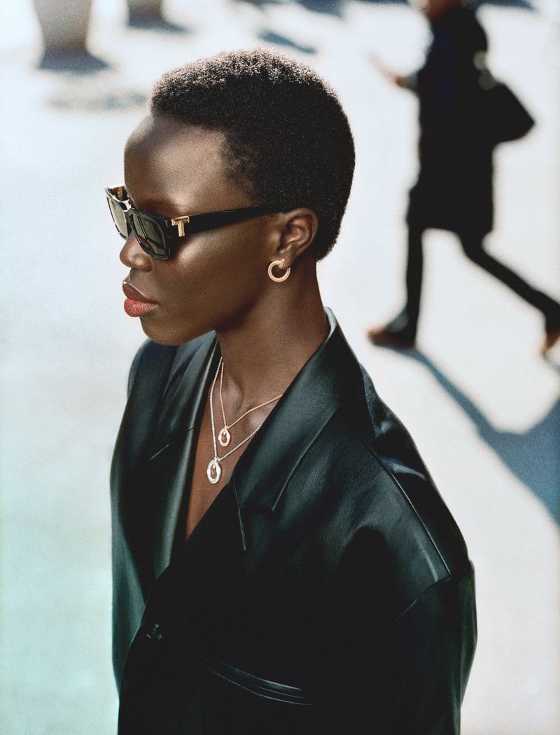 Model wearing dark sunglasses, gold hoop earrings, layered necklaces, and a black leather jacket against an urban street background.