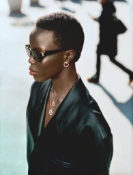 Model wearing dark sunglasses, gold hoop earrings, layered necklaces, and a black leather jacket against an urban street background.
