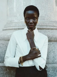Close-up portrait of a Black woman wearing a white collared shirt and gold jewelry against a stone architectural pillar background
