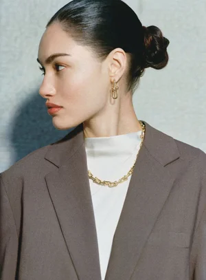 Model wearing a brown blazer, white top, and gold jewellery with hair in a bun against a neutral textured wall.