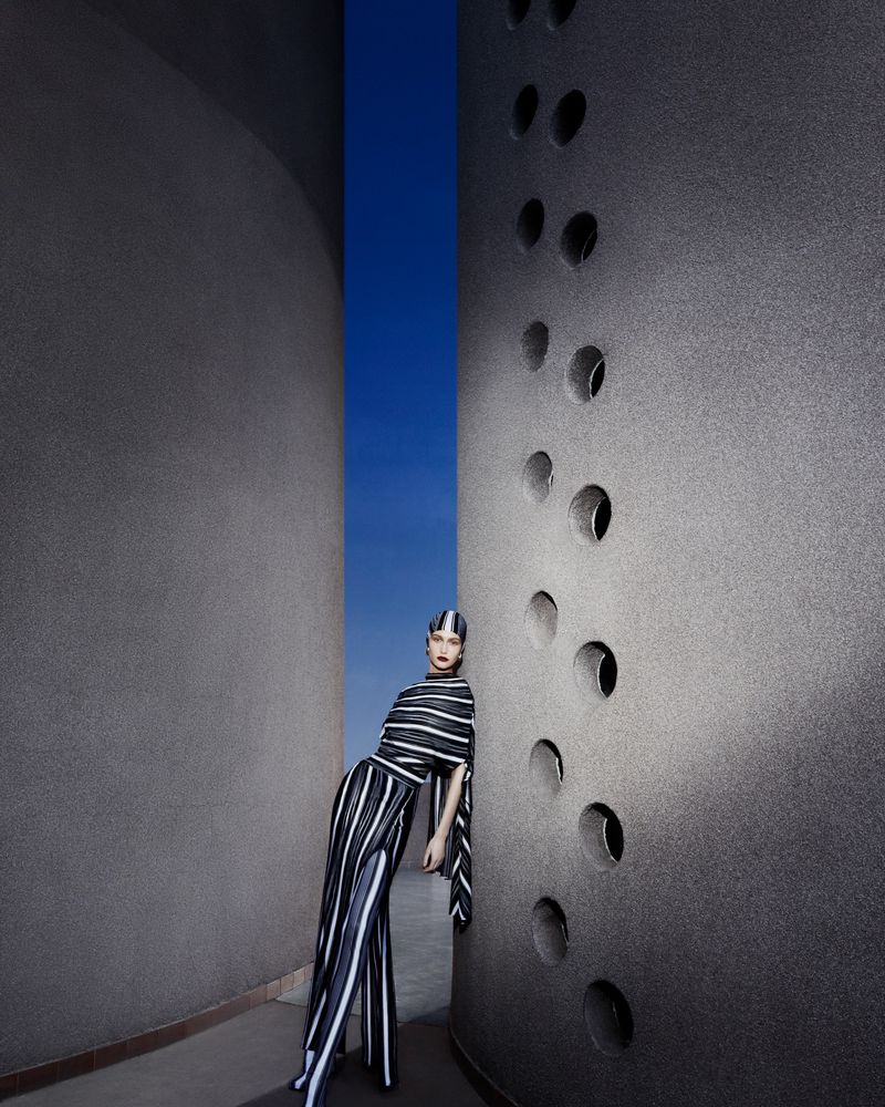 Fashion model in striped top and pants leaning against a textured wall with circular cutouts against a blue sky backdrop.
