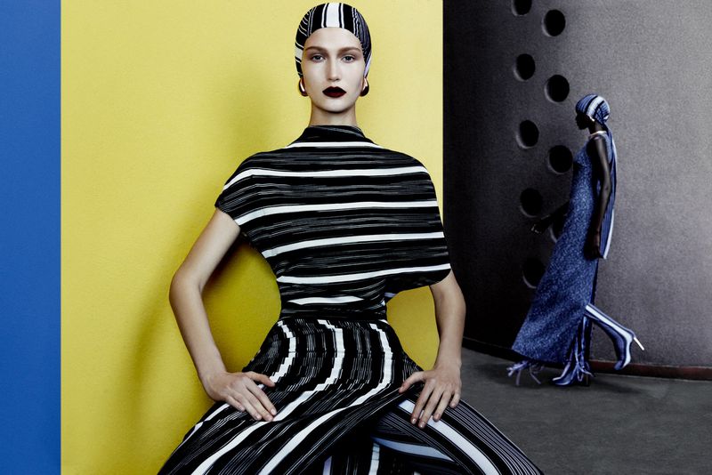 Fashion shot of an adult female model in a striped outfit seated against a yellow background, another model standing in the background.