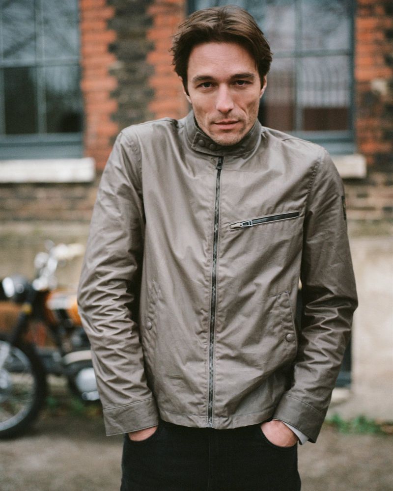 Stylish adult male in grey jacket posing outdoors, with vintage motorbike parked in background, neutral color palette, medium shot.