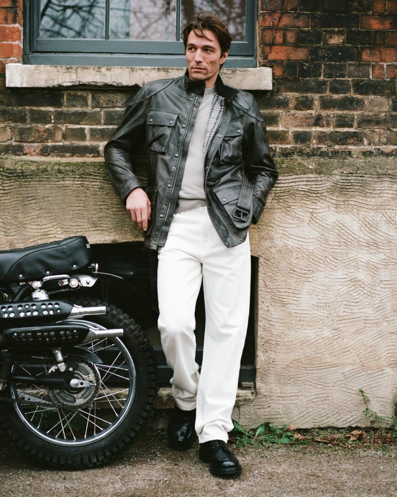 Model leaning against a weathered brick wall next to a vintage motorcycle wearing a dark leather jacket and white trousers