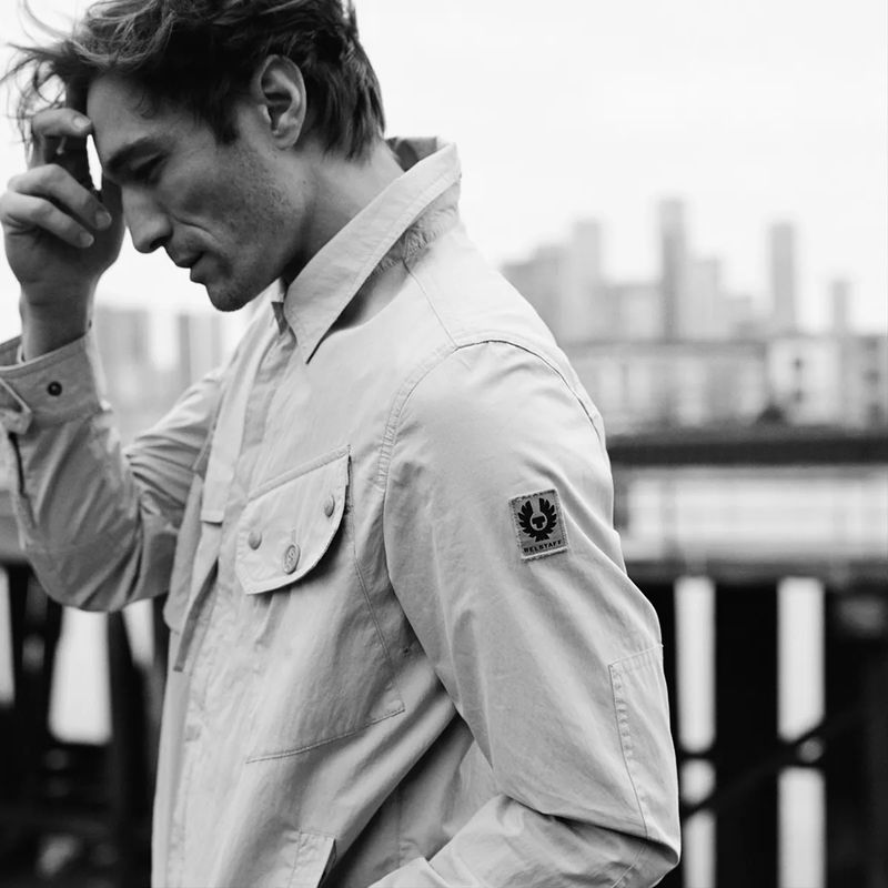 A man with dark hair wearing a light-colored jacket stands outdoors, gazing downward with a blurred city skyline in the background.