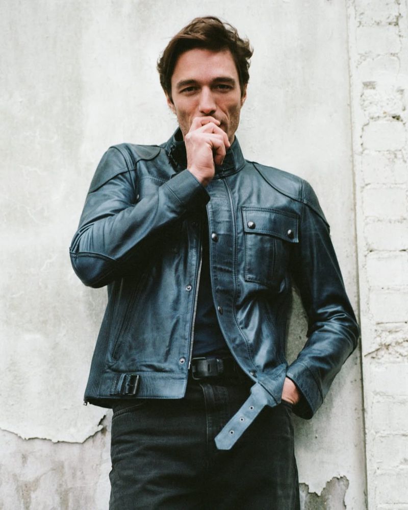 A handsome adult man in a leather jacket poses indoors against a concrete wall. The lighting is soft and diffused.