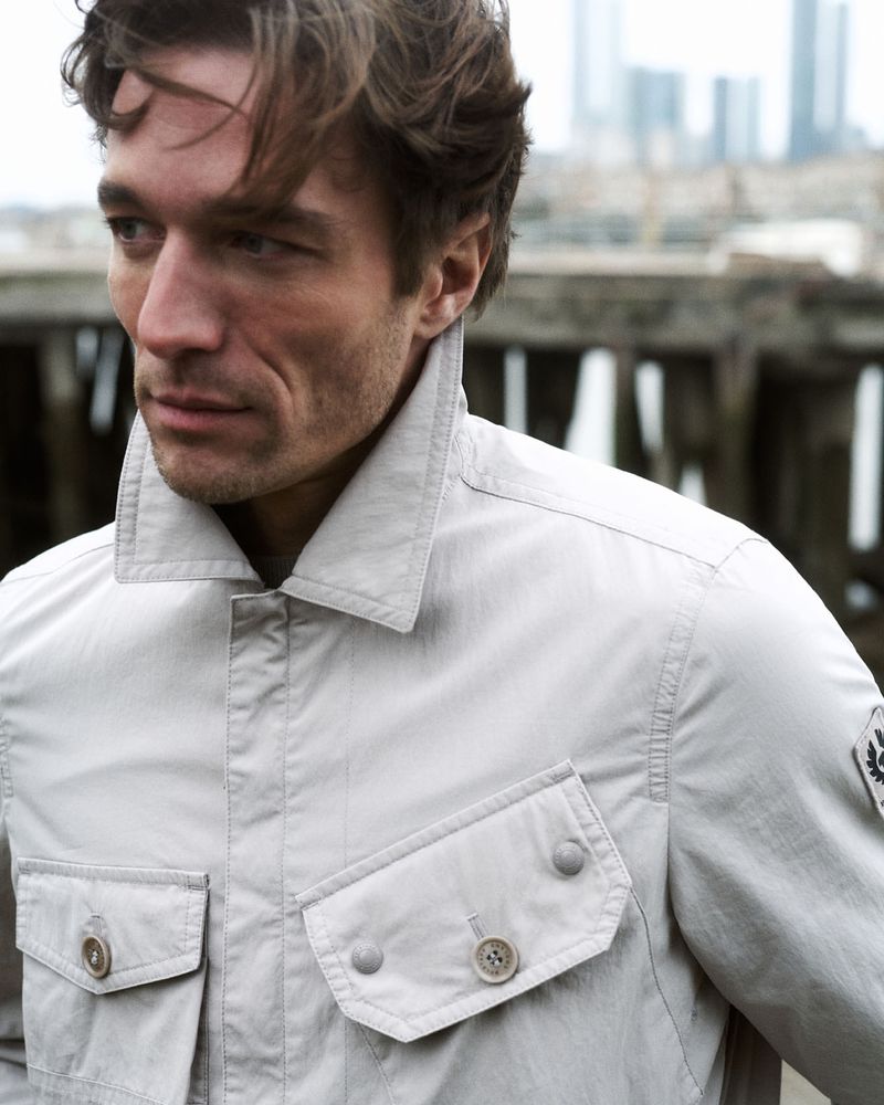 Close-up of adult male in gray jacket posing outdoors against an urban waterfront background with a blurred cityscape in the distance.
