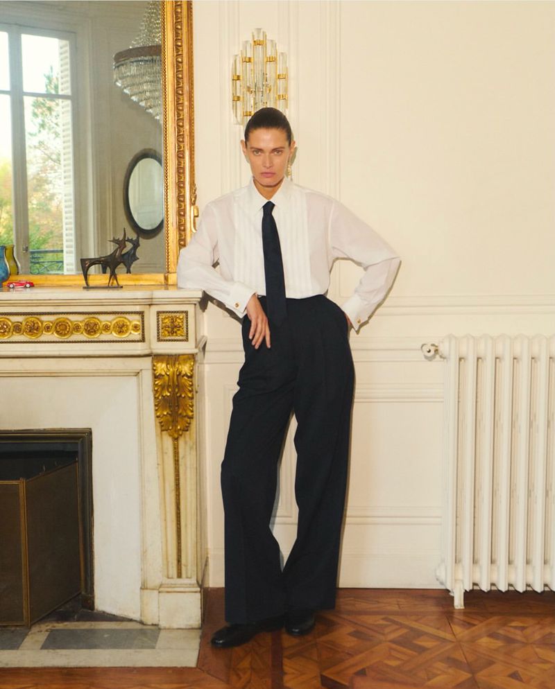 Adult woman in a white button-down shirt and black tie and pants, leaning on a fireplace, looking at the camera in an indoor setting.