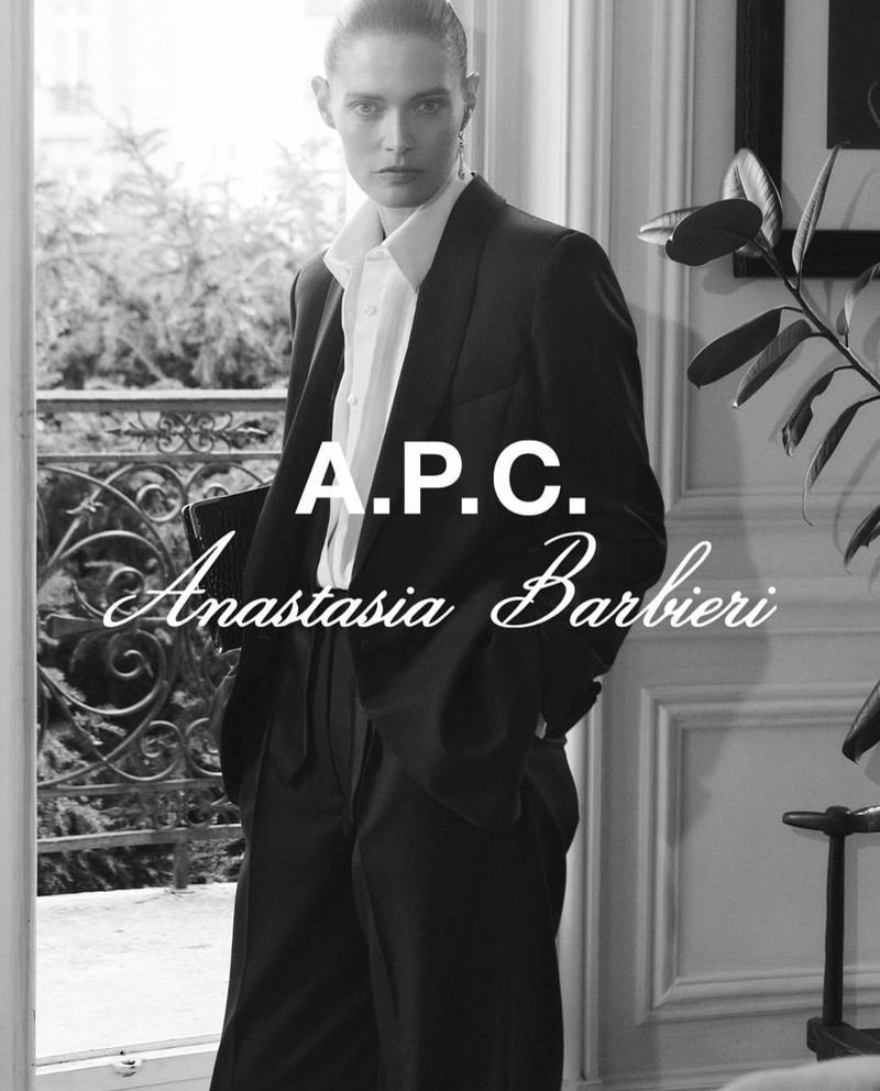 A monochromatic portrait of a female model wearing a black suit jacket and white button-down shirt, gazing intently at the camera indoors.