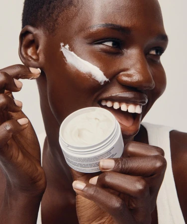 Close-up of a smiling Black woman holding a jar of moisturizer with a white cream application on her cheek in a studio setting.