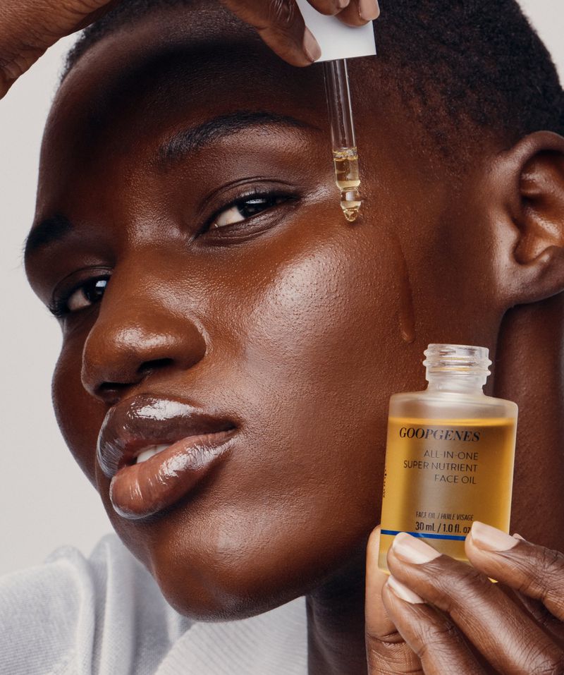 Close-up of a dark-skinned woman's face with a dropper applying oil to her cheek, holding a bottle of face oil. Beauty shot.