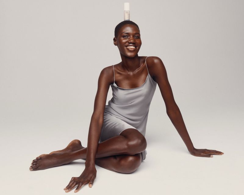 Woman poses while balancing cosmetic bottle on head in studio with a soft smile. She wears a gray dress, sitting with arms resting.