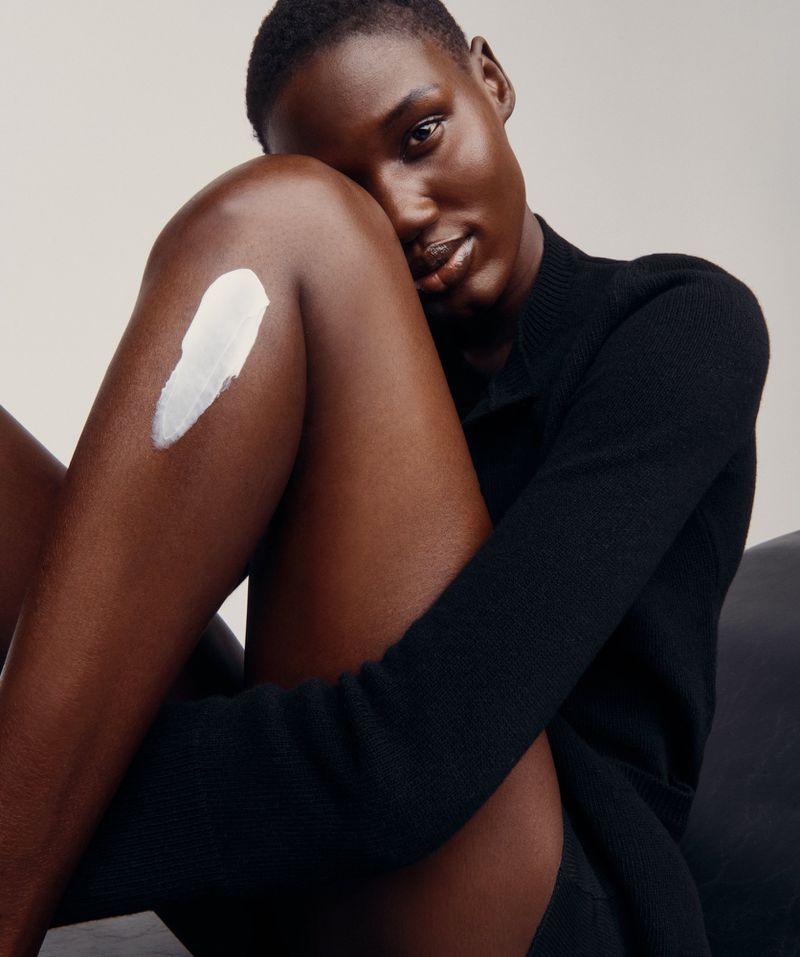 Portrait of a dark-skinned woman in a black top, seated and posing with cream on her thigh, set against a plain light backdrop.