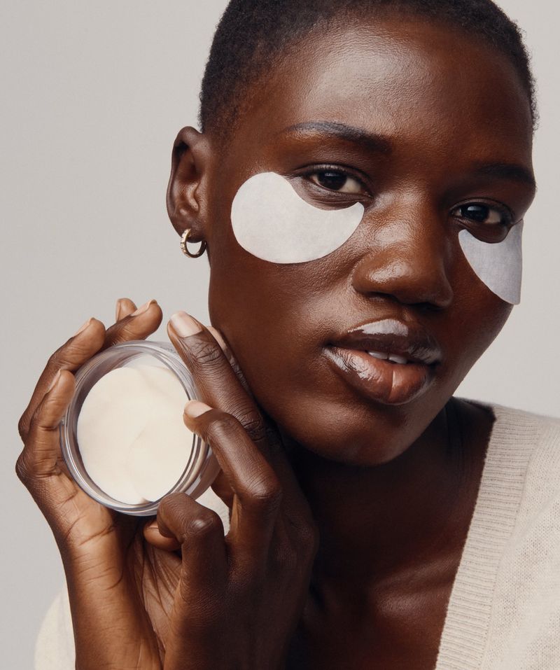 Close-up portrait of an African woman with eye patches, holding a skincare product in a jar. Beauty and skincare concept.