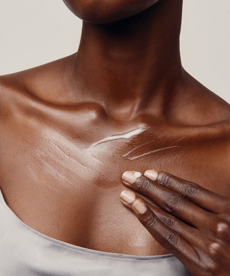 Close-up of an African American adult female applying white lotion to her chest indoors, with a neutral background.