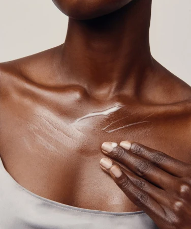 Close-up of an African American adult female applying white lotion to her chest indoors, with a neutral background.