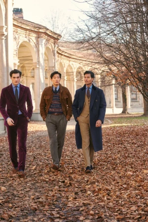 Three stylish men in suits walk on a path covered in autumn leaves near an arcade with arched openings, showcasing a blend of fashion and architecture.