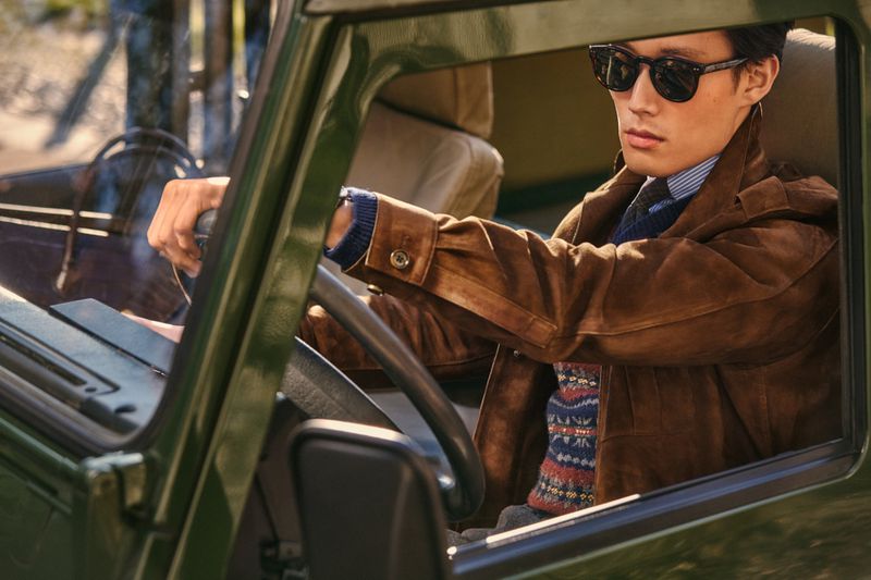 Man driving an olive green car, wearing a brown suede jacket and sunglasses. The atmosphere is vintage with an outdoor setting.