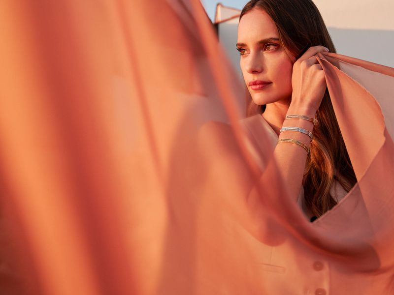 Woman with long brown hair wearing bracelets and a light pink top, standing outside as the sun sets, soft, airy aesthetic.