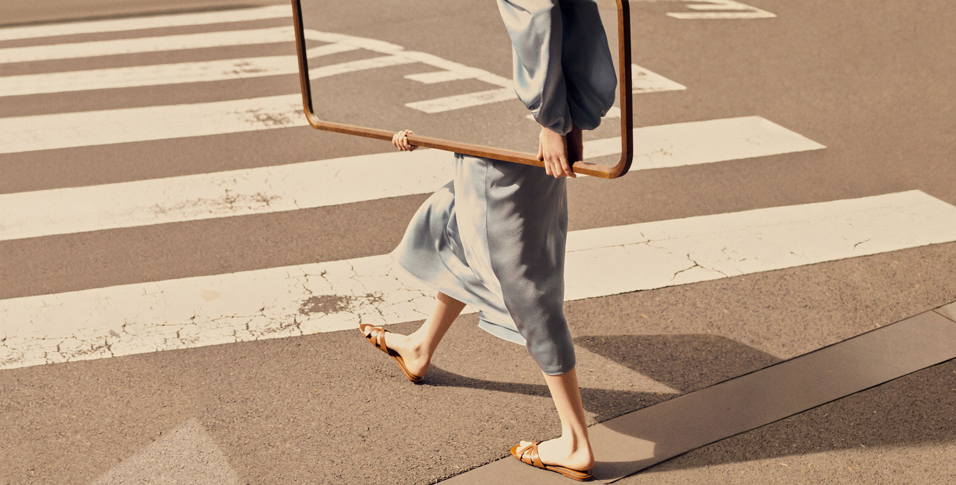 A woman in a blue dress and sandals walks across a crosswalk holding a large, square frame on a city street during daylight.