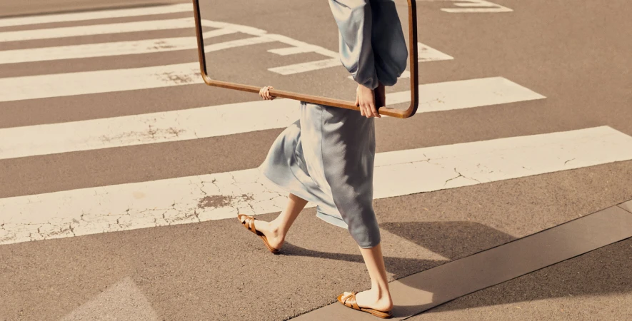 A woman in a blue dress and sandals walks across a crosswalk holding a large, square frame on a city street during daylight.