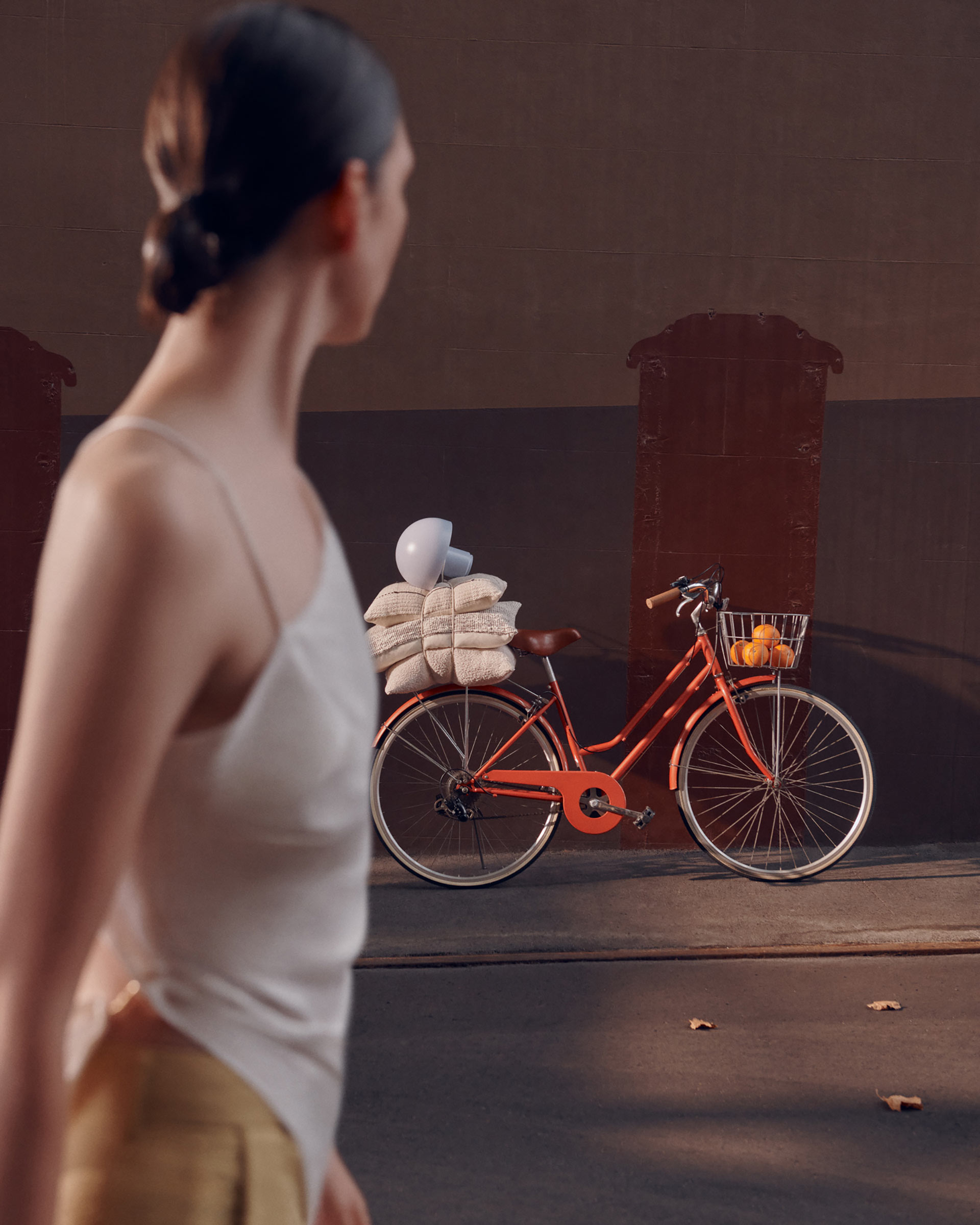 Woman in a slip dress walking by a red bicycle with a basket of oranges and luggage in an urban outdoor environment.