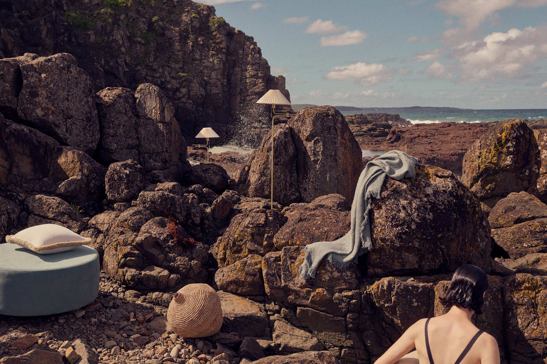 Coastal scene with a woman, lamps, blanket, rocks, and ocean. Natural setting with interior design elements in outdoor lifestyle photo.