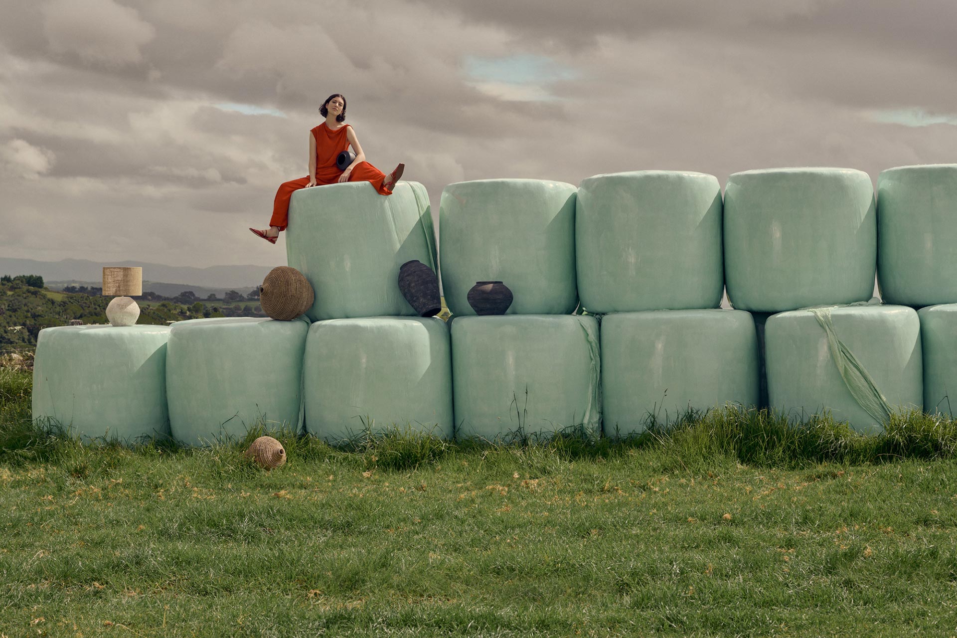 Woman in an orange jumpsuit sits atop a stack of wrapped hay bales in a grassy field under a cloudy sky. Pottery displayed on bales.
