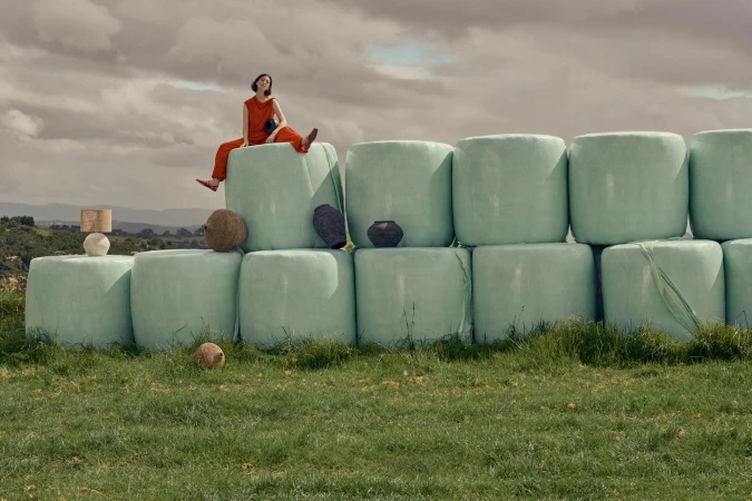 Woman in an orange jumpsuit sits atop a stack of wrapped hay bales in a grassy field under a cloudy sky. Pottery displayed on bales.