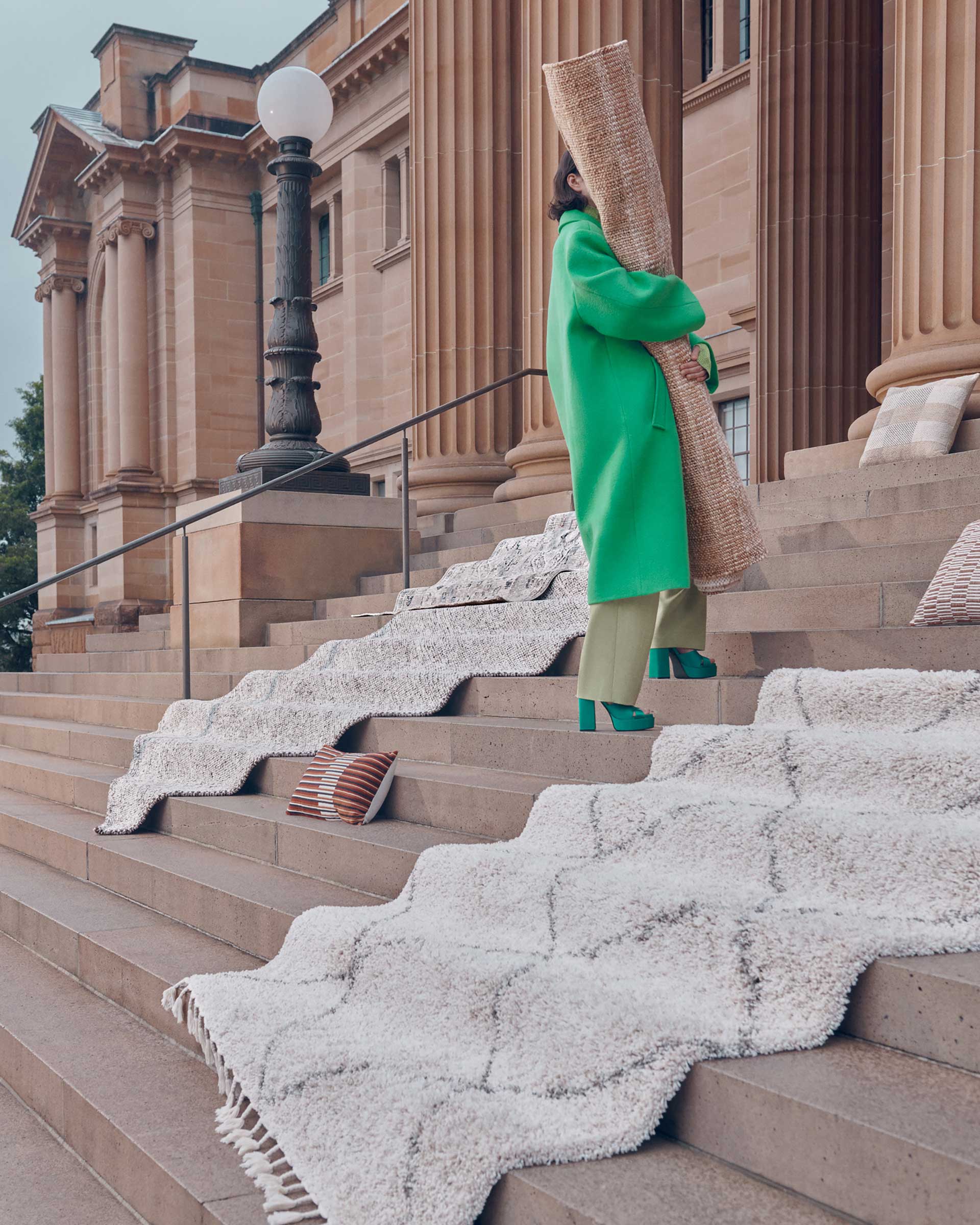 Woman in green coat carrying rolled carpet on stairs with a beige patterned carpet, a pillar building and overcast sky in background.