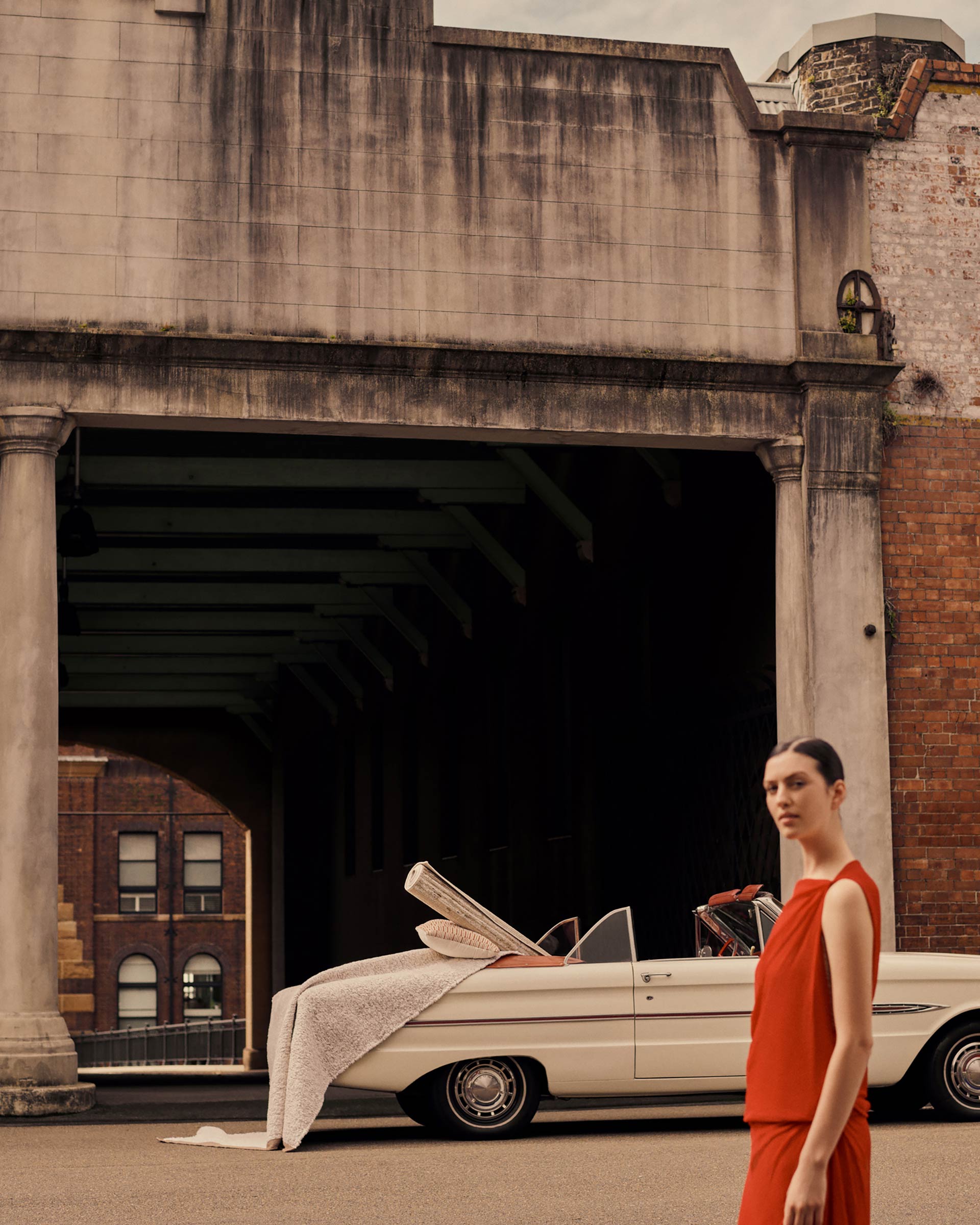 Woman in red dress stands near a vintage car under a brick tunnel, creating a fashion editorial shot with urban, outdoor environment.