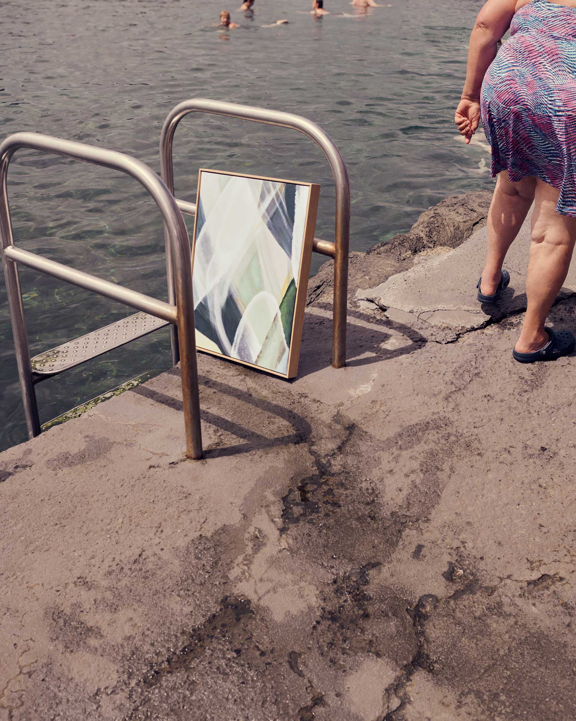 A framed abstract artwork is propped against a metal railing near the water's edge, with swimmers and a woman in the background.