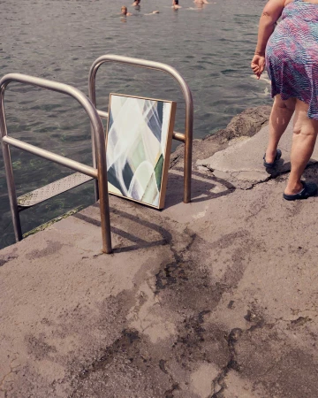 A framed abstract artwork is propped against a metal railing near the water's edge, with swimmers and a woman in the background.