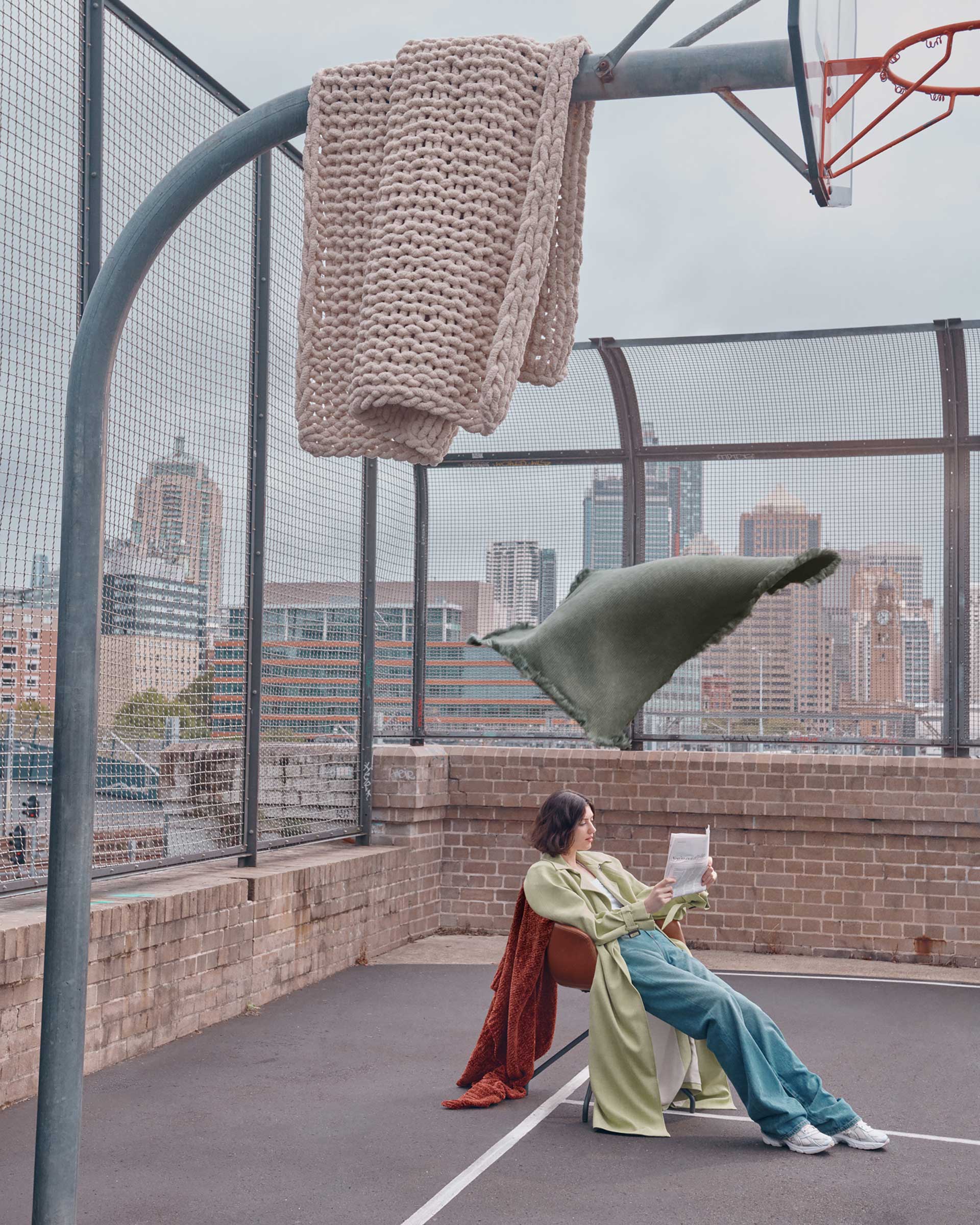 Woman sits in an outdoor basketball court reading, with floating blankets and cityscape in the background, under overcast lighting.