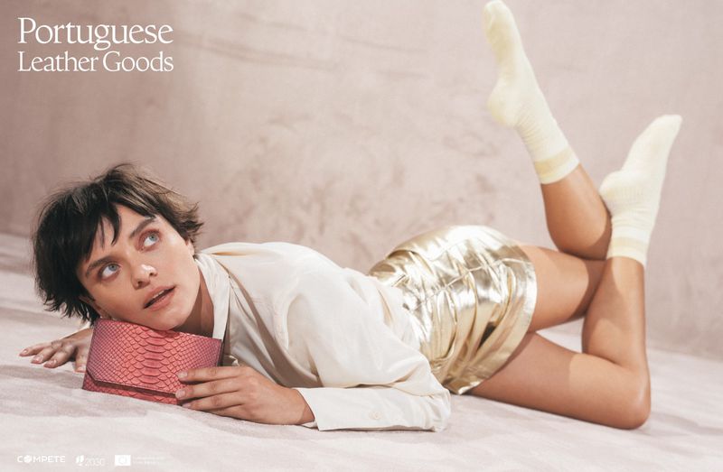 Woman lying down with handbag, wearing gold shorts and socks, and a light button-down shirt against a pink textured backdrop.