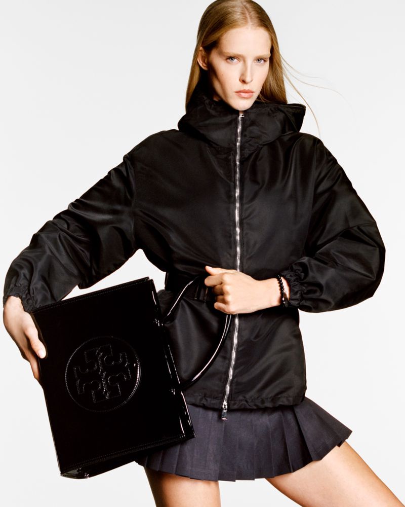 Model Abby Champion wearing a black jacket and holding a black patent leather handbag against a white background for Tory Burch S/S 2025.