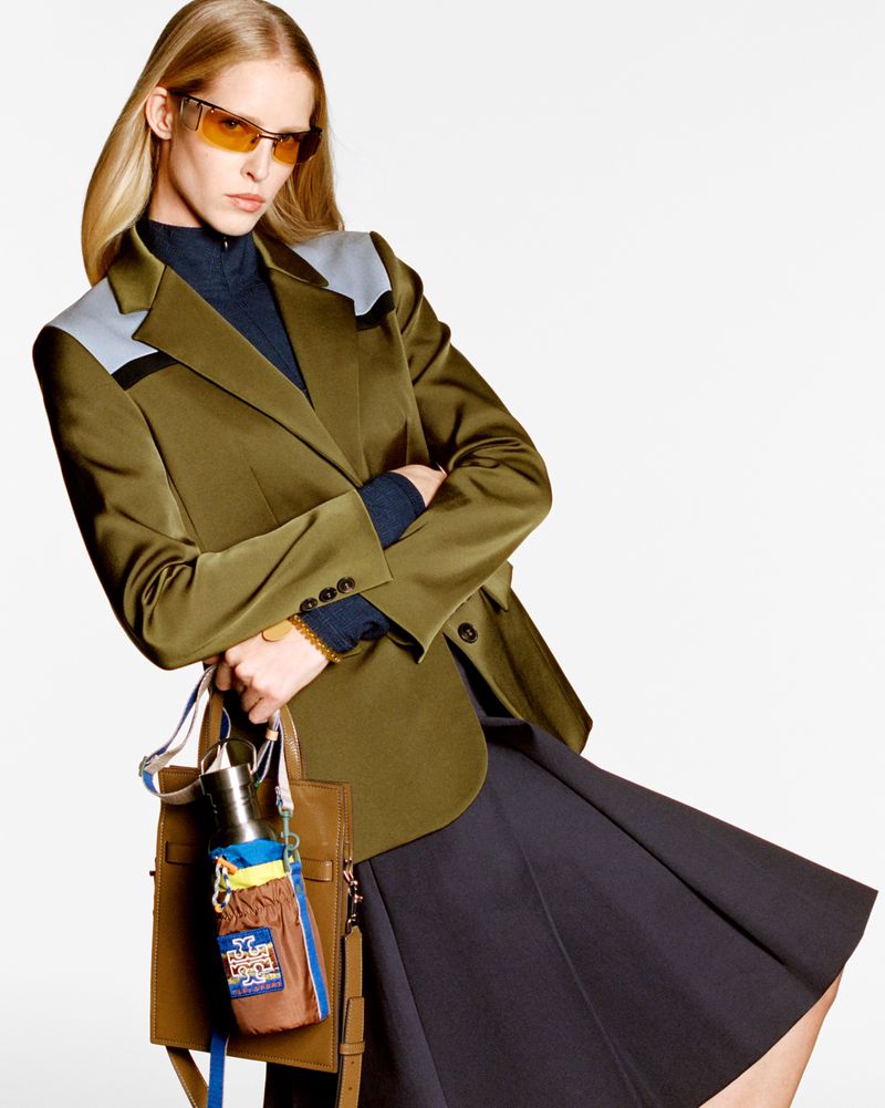 Adult female model in a green blazer, sunglasses, and black skirt poses in a studio holding a brown tote bag with a water bottle.