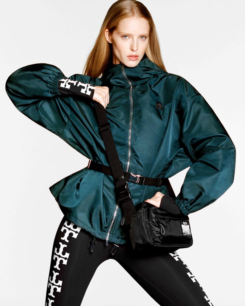 Blonde woman poses in a dark green hooded jacket with black leggings and crossbody bag against a white backdrop in a fashion shot.