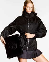 Model Abby Champion wearing a black jacket and holding a black patent leather handbag against a white background for Tory Burch S/S 2025.
