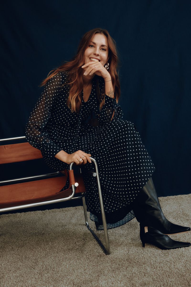 A woman with long brown hair sits in a chair smiling. She wears a navy dress with white polka dots and black heeled boots.