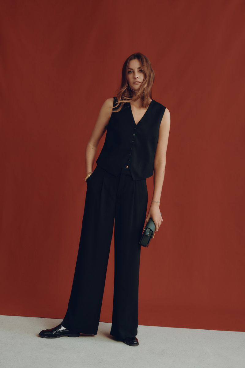 A woman in a sleeveless black vest and black trousers stands against a terracotta background holding a clutch, showcasing a modern, sleek style.