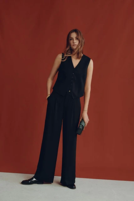 A woman in a sleeveless black vest and black trousers stands against a terracotta background holding a clutch, showcasing a modern, sleek style.