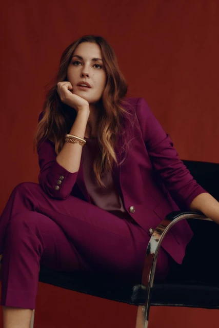 Portrait of a woman with long brown hair in a magenta pantsuit sitting in a black chair against a reddish-brown backdrop in a studio.
