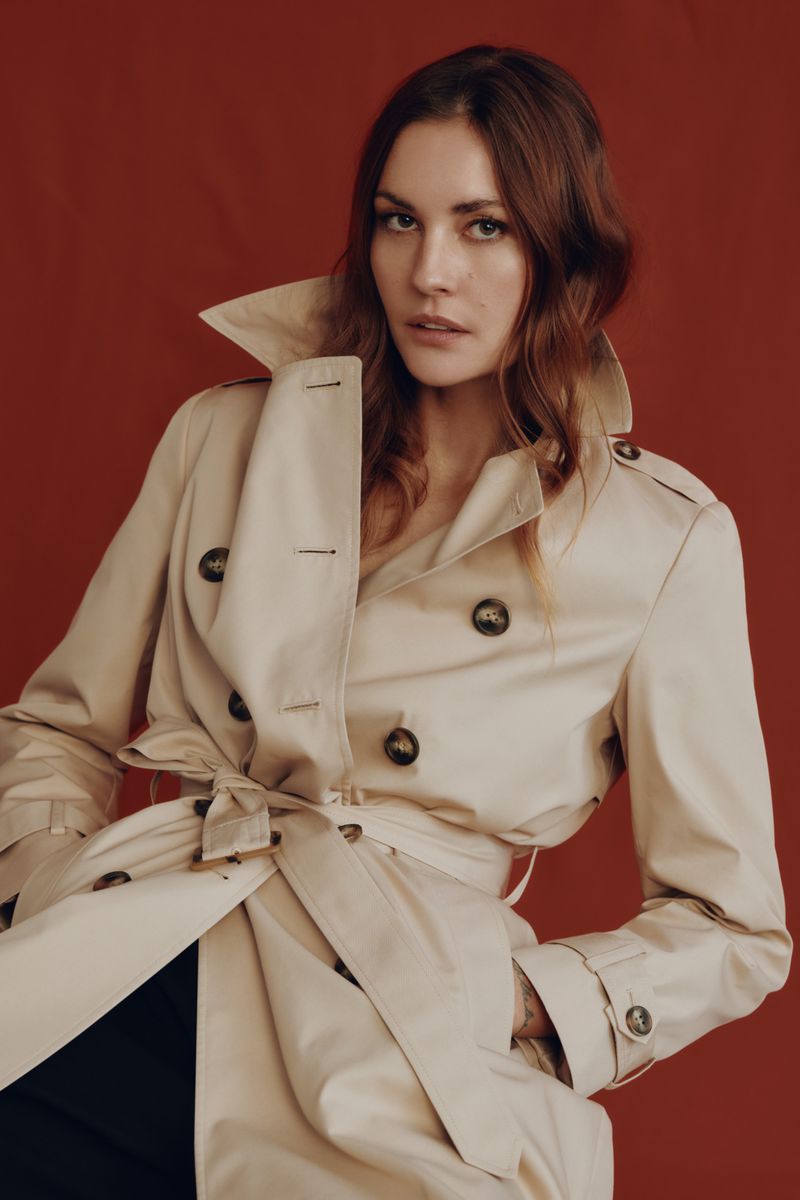 Close-up shot of a woman in a beige trench coat against a red background in a fashion portrait, showcasing classic outerwear.