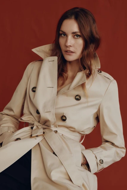 Close-up shot of a woman in a beige trench coat against a red background in a fashion portrait, showcasing classic outerwear.