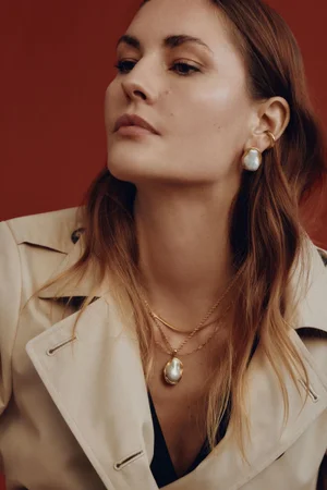 Model Nadja Bender wearing pearl earrings, layered gold necklaces, and a beige trench coat against a warm rust-red background.